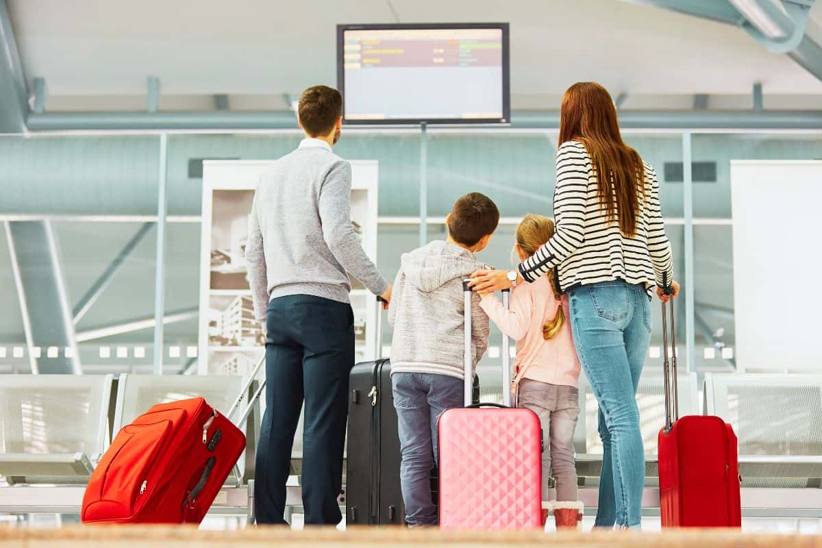 A family of mum, dad and 2 children standing in an airport with suitcases looking at a screen for travel information.